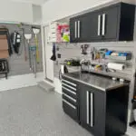 A well-organized garage features a sleek black storage cabinet with multiple drawers and shelves, a workbench with various tools and supplies, wall-mounted racks holding cleaning tools and sports equipment, and a pristine, speckled gray floor.