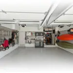 A well-organized garage with two separate parking spaces. The left side features organized shelves and hooks holding tools, camping gear, and bicycles. The right side has wall-mounted storage for two kayaks. A workbench with cabinets is in the center back wall.