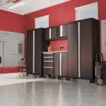 A well-organized garage with red walls features black storage cabinets, a stainless steel countertop, and tools neatly hung on wall panels like a template of efficiency. Sunlight streams through two small windows above the cabinets, illuminating the clean, spacious floor.